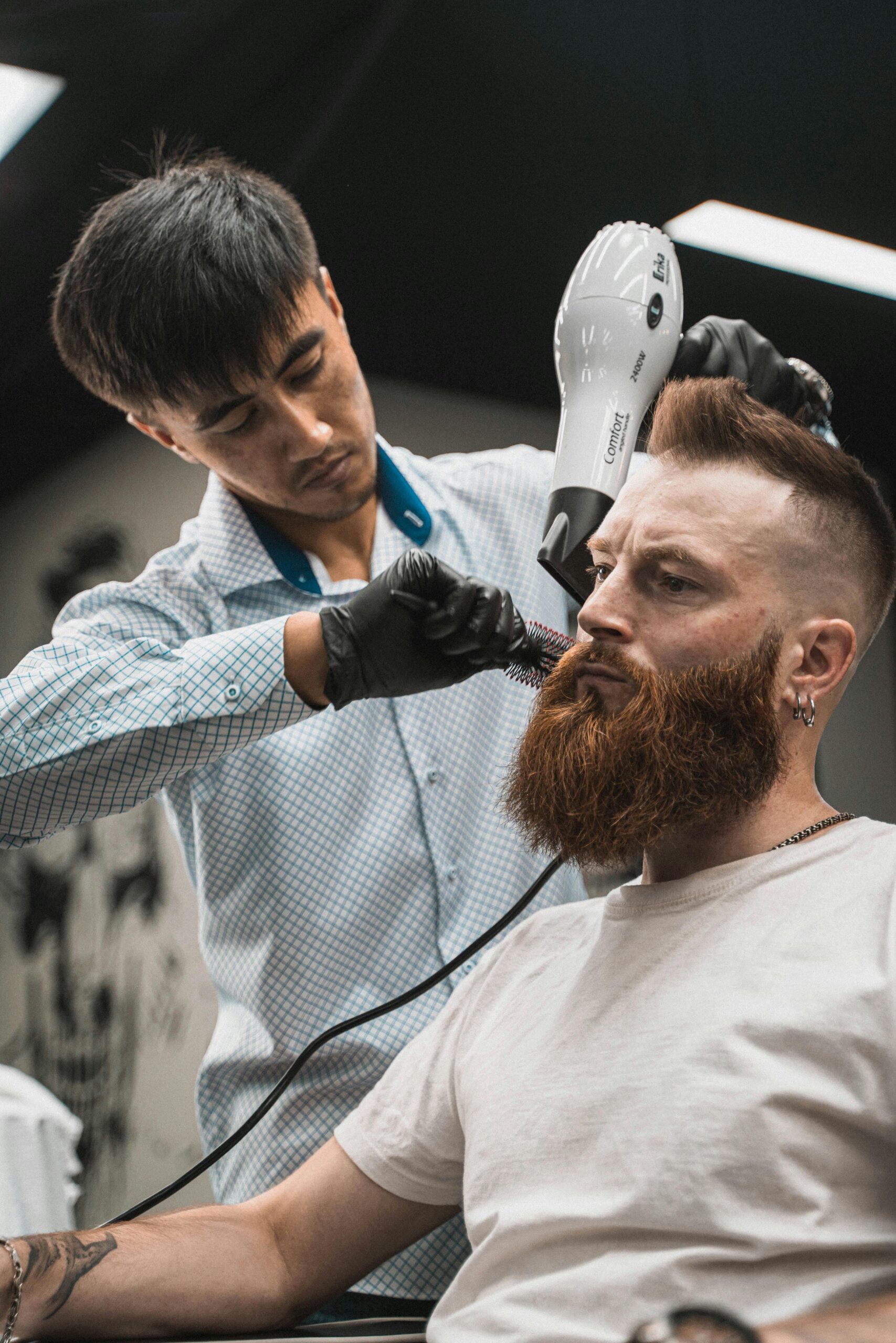 Barber styling a bearded man's hair with a blow dryer in a modern barbershop.