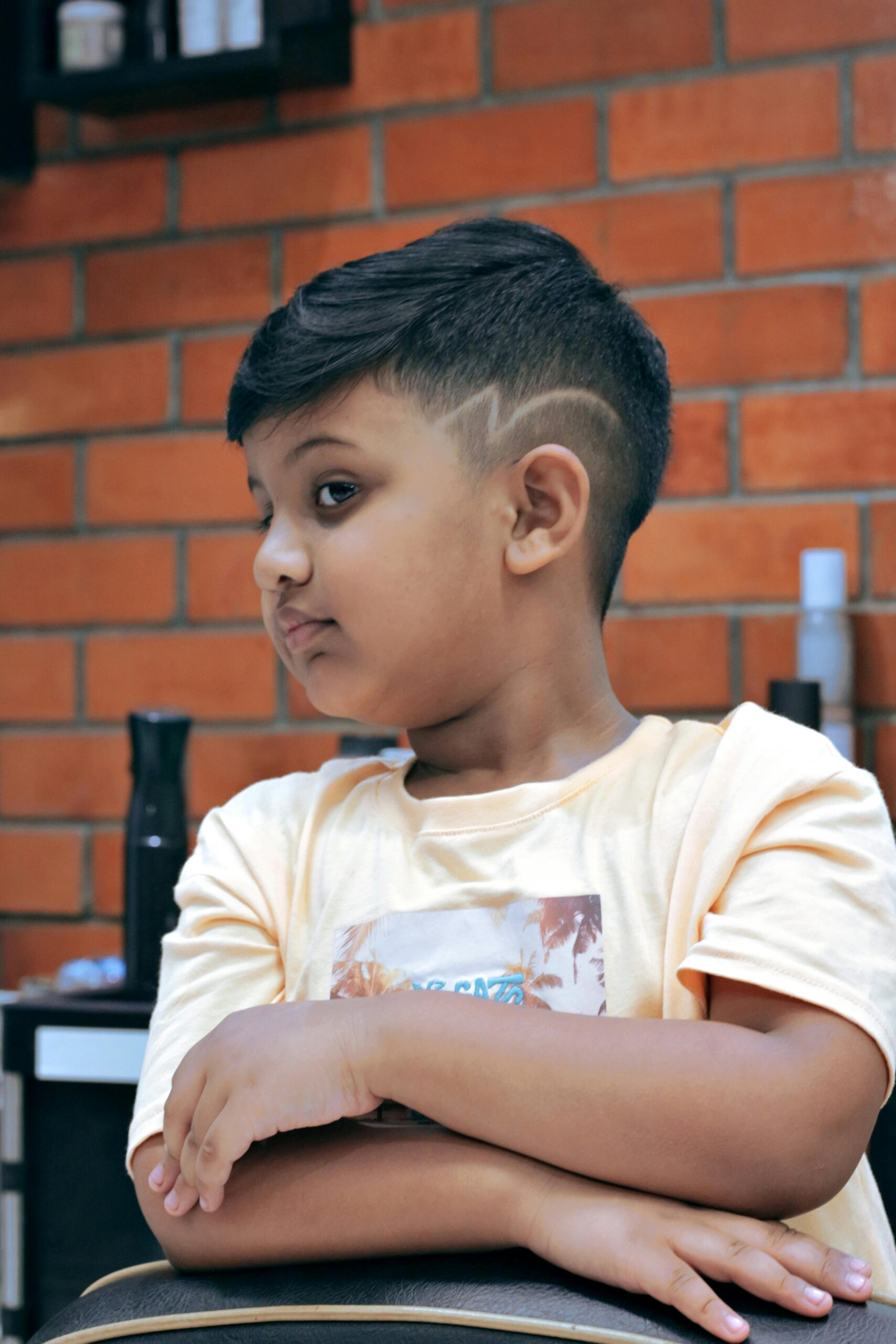 Child sitting in a barbershop showcasing a trendy hairstyle against a brick wall background.